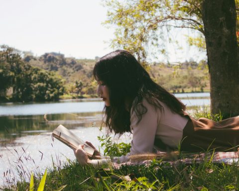 woman reading book and lying forward on sheet on grass beside body of water during day