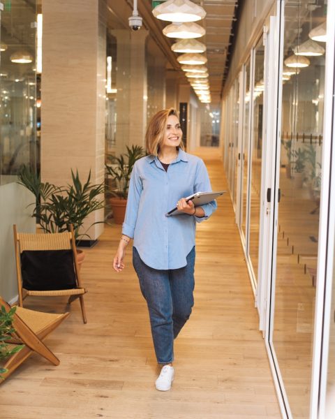 woman in blue dress shirt and blue denim jeans standing beside brown wooden chair