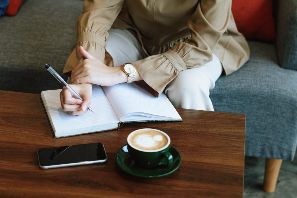 Jonge vrouw schrijft in een notitieboekje met een kop koffie naast zich, bezig met zelfreflectie